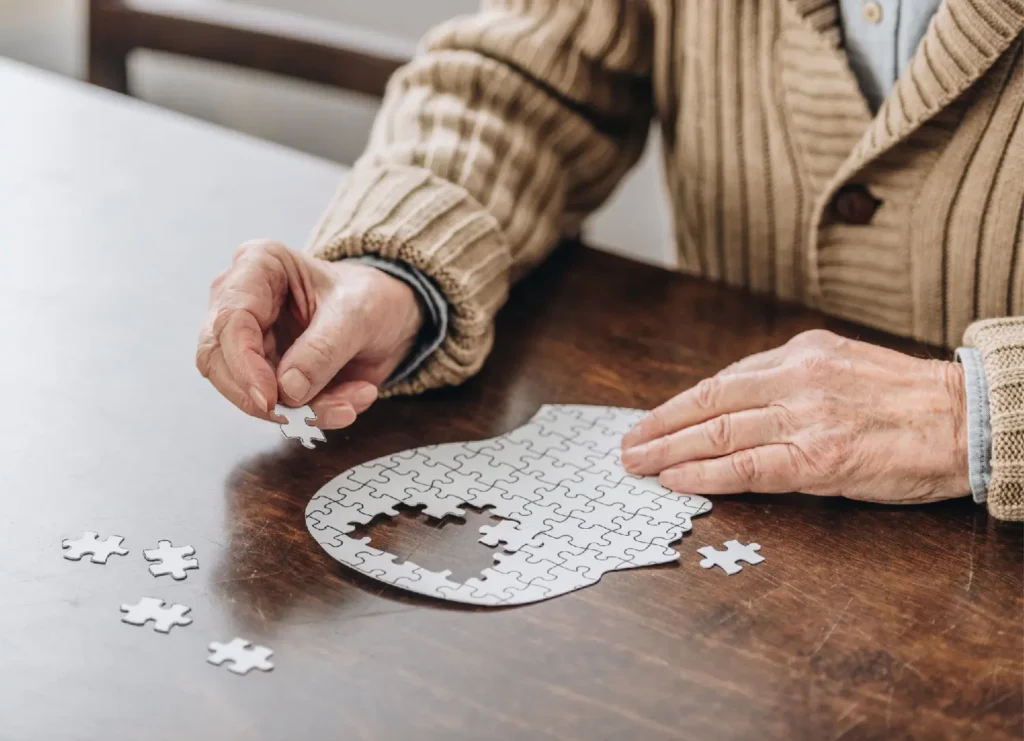 Zbliżenie na dłonie starszej osoby układającej białe puzzle na drewnianym stole; układanka ma kształt ludzkiej głowy z brakującymi elementami w środku