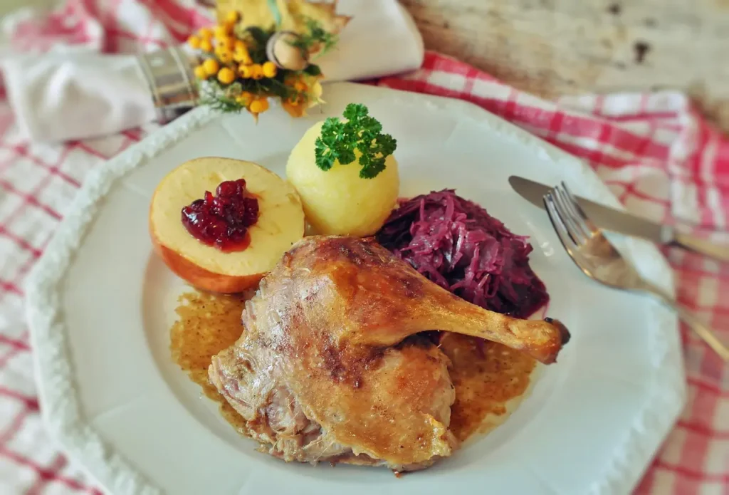 Apetyczne danie obiadowe na białym talerzu: pieczone udo kaczki w sosie, podane z czerwoną kapustą, knedlem ziemniaczanym oraz pieczonym jabłkiem z żurawiną