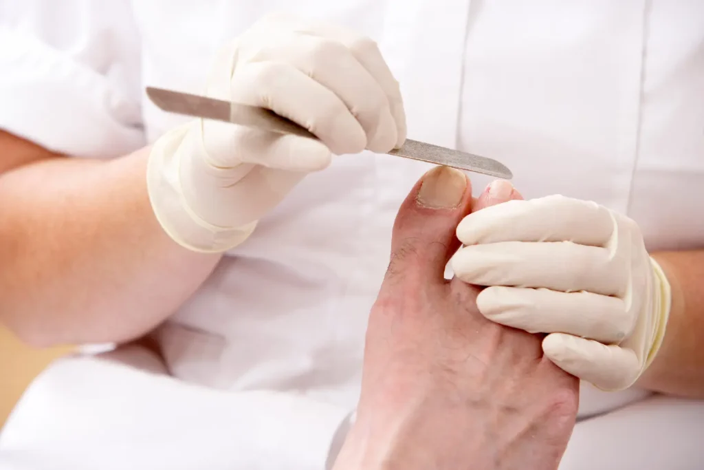 Zbliżenie na dłonie w białych rękawiczkach wykonujące zabieg pedicure (piłowanie paznokcia u stopy) u starszej osoby