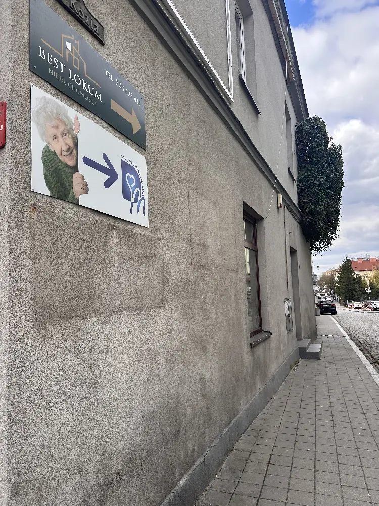 Zdjęcie narożnika szarej kamienicy przy chodniku na ulicy Koziej. Na ścianie wiszą dwa szyldy: czarny z napisem "BEST LOKUM NIERUCHOMOŚCI" oraz biały z wizerunkiem uśmiechniętej starszej pani oraz logiem Domowej Opieki Mobilnej, prowadzący do wejścia biura