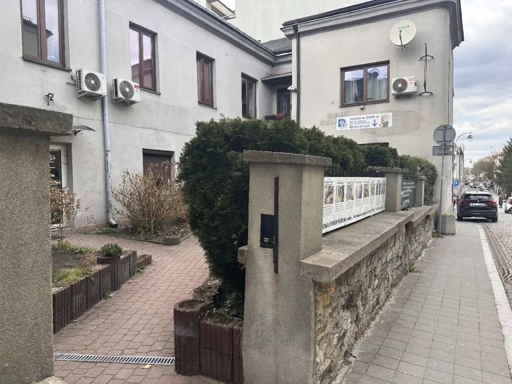 Perspektywa z poziomu chodnika na podwórze biurowe na ulicy Koziej. Widoczny kamienny murek z banerem reklamowym Domowej Opieki Mobilnej, gęsty zielony krzew i elewacja budynku oraz szyld z napisem "Jesteś w DOM-u"