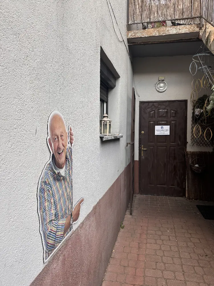 Fragment elewacji obok brązowych drzwi. Na ścianie umieszczona jest wycięta tablica reklamowa w kształcie uśmiechniętego starszego mężczyzny wskazującego palcem w stronę wejścia do biura