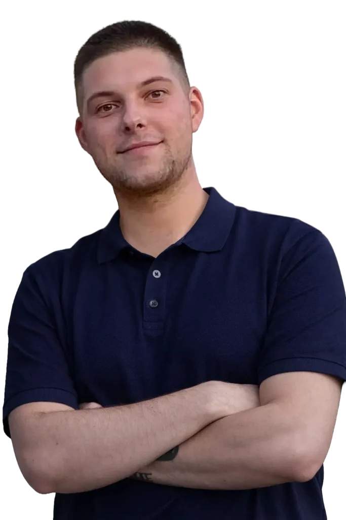 Portret uśmiechniętego młodego mężczyzny w ciemnogranatowej koszuli, z krótkimi ciemnymi włosami, stojącego z założonymi rękami