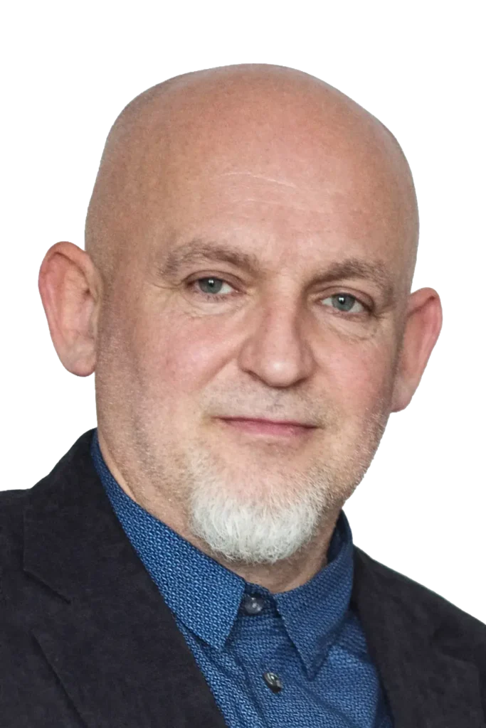Portret uśmiechniętego mężczyzny w ciemnej marynarce i niebieskiej koszuli, z krótką siwą brodą i ogoloną głową