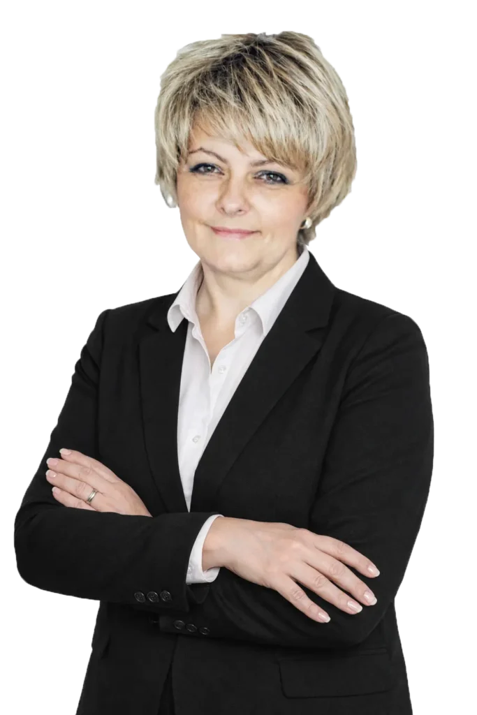 Portret uśmiechniętej kobiety w ciemnej marynarce i białej koszuli, z krótkimi blond włosami, stojącej z założonymi rękami