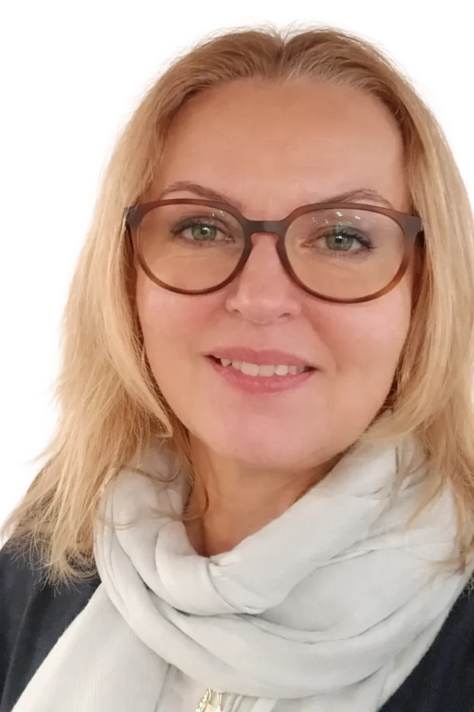 Portret uśmiechniętej kobiety w ciemnym płaszczu i białym szalu, z długimi blond włosami i okularami